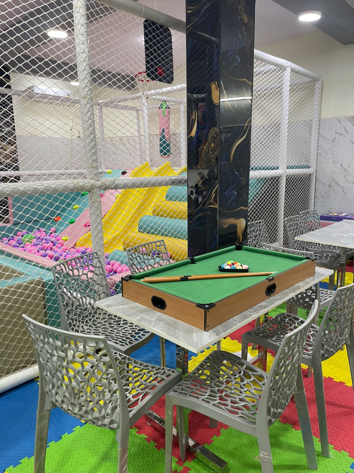 A mini pool table sits on a table in a brightly colored indoor playground with a ball pit and slides.