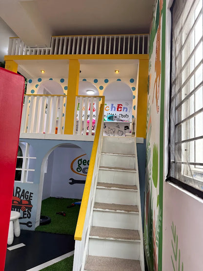 A colorful indoor playhouse with a kitchen and garage theme, featuring a staircase and playful decor.