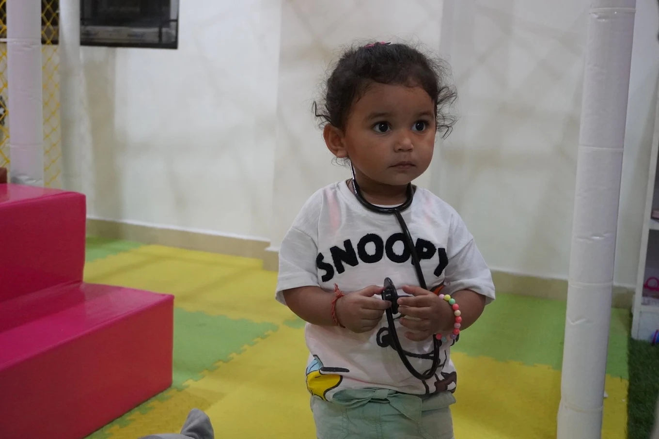 A young child dressed as a doctor plays with a stethoscope in a colorful indoor play area.