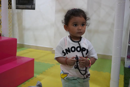 A young child dressed as a doctor plays with a stethoscope in a colorful indoor play area.
