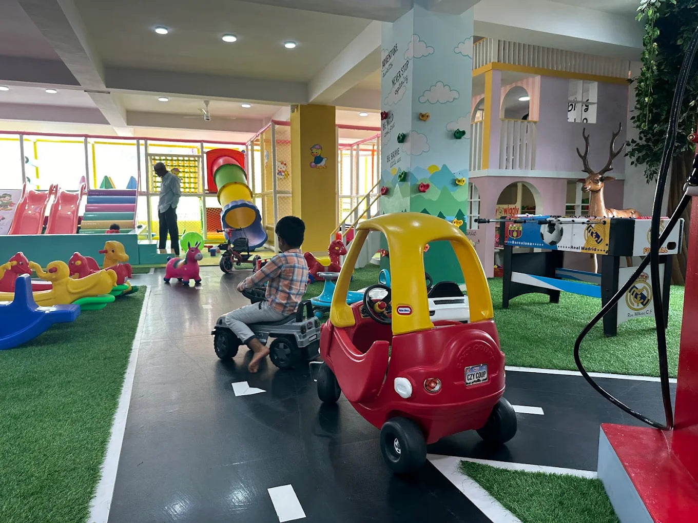 Children play in a colorful indoor playground with slides, ride-on toys, and a foosball table.
