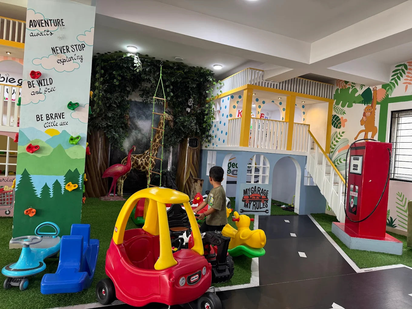 A young boy plays in a colorful indoor playground with a climbing wall, toy cars, and a playhouse.