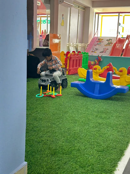 A young boy plays with a toy jeep and ring toss on artificial grass in a colorful indoor play area.