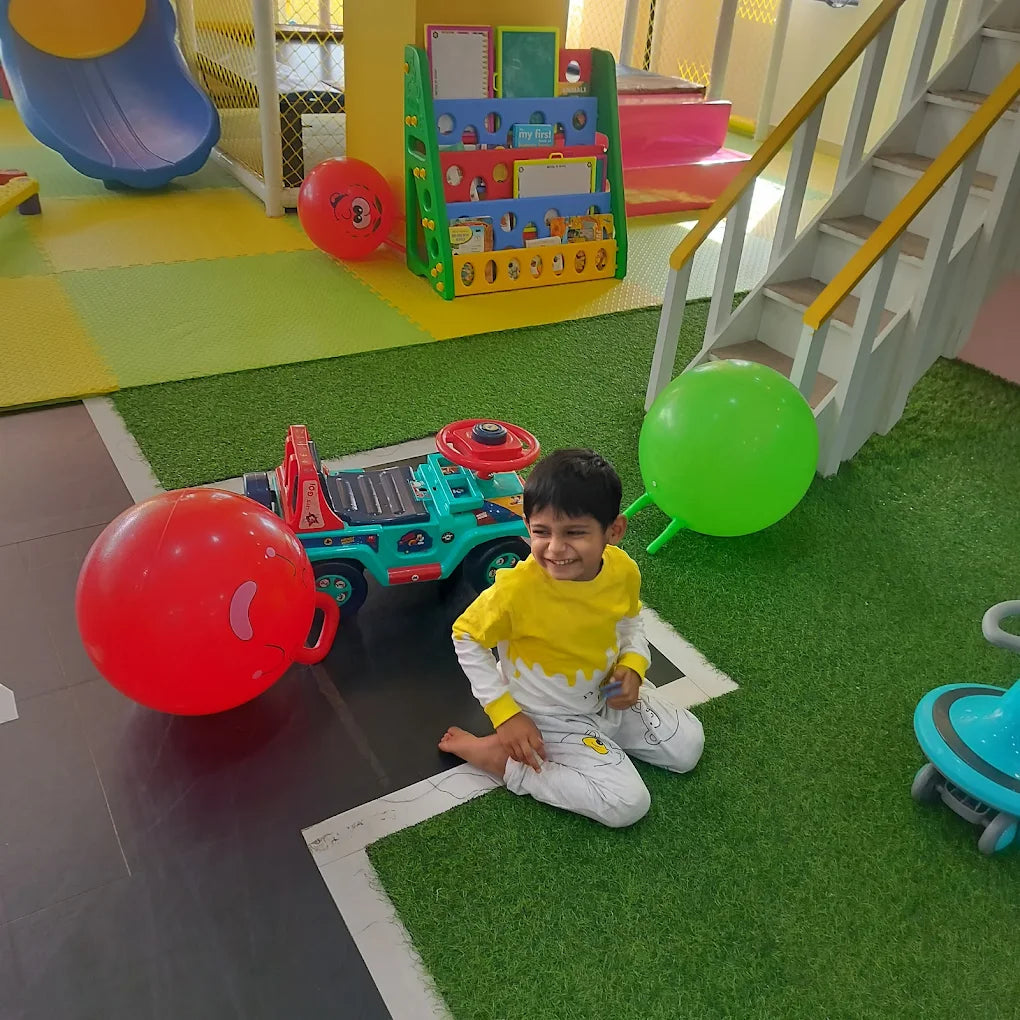 A young boy sits on a green carpet in a play area, smiling next to a toy car and large red and green balls.