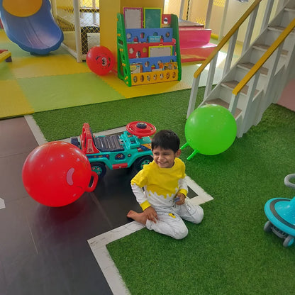 A young boy sits on a green carpet in a play area, smiling next to a toy car and large red and green balls.