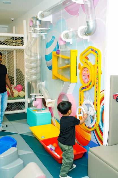 A young boy plays with a colorful ball-dropping machine in a bright, indoor play area.