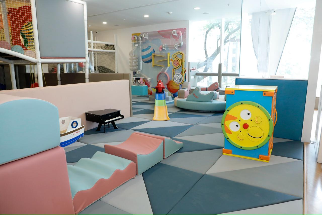 A colorful indoor play area for young children with soft play structures and a wall activity panel.