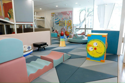 A colorful indoor play area for young children with soft play structures and a wall activity panel.