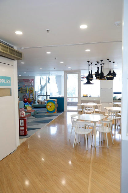 A bright, modern play area with tables and chairs for children, featuring a colorful play structure and decorative gas pumps.