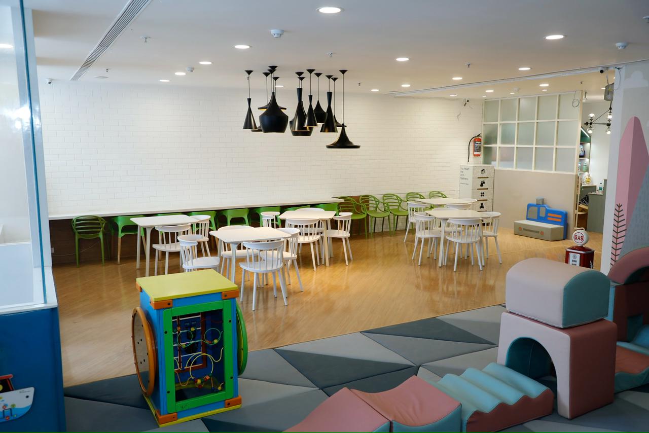 A brightly lit indoor play area with tables, chairs, and soft play structures for children.