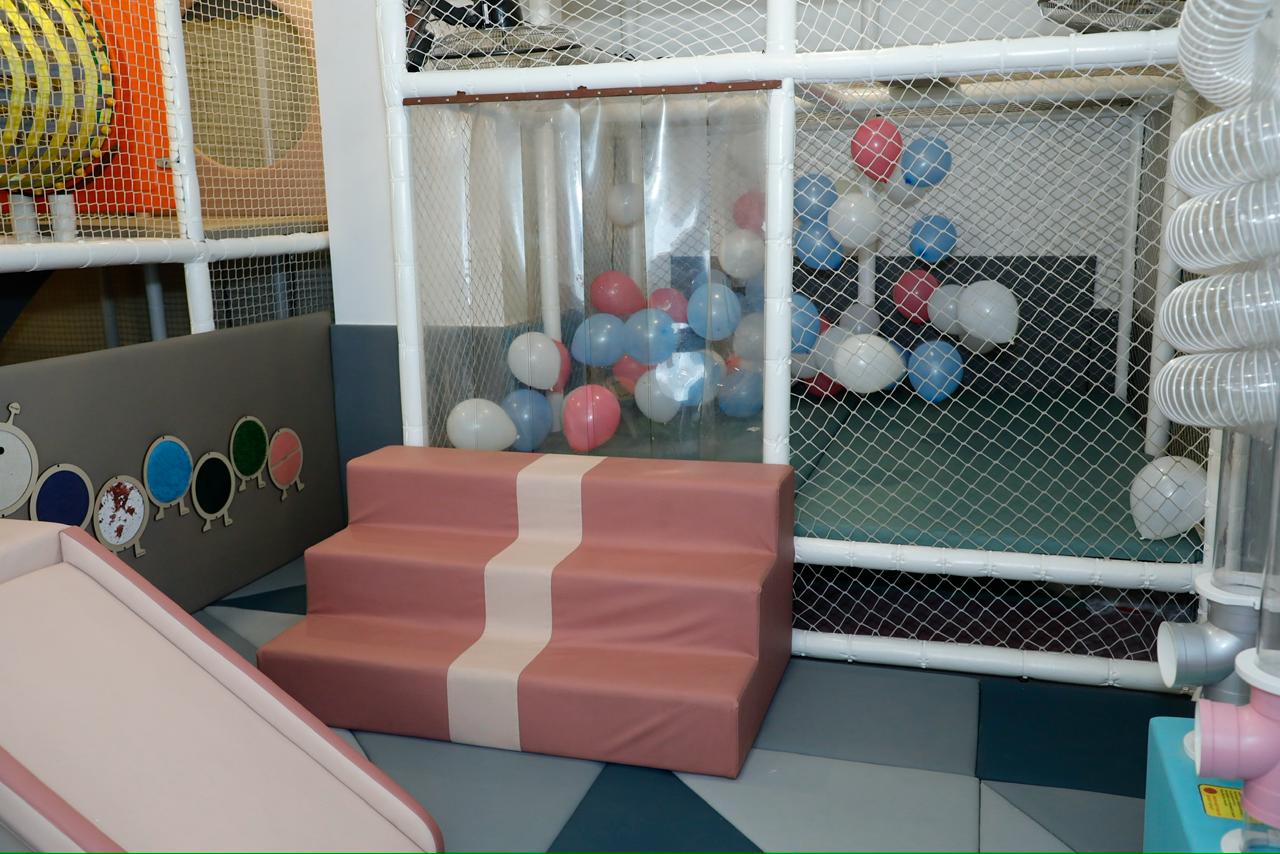 A soft play area for children with a pink slide, a tiered pink step, and a ball pit filled with balloons.