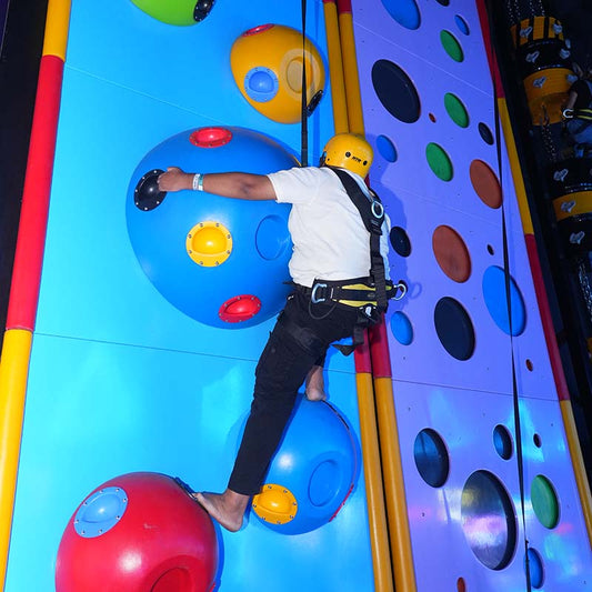 A person in a harness climbs a colorful indoor rock-climbing wall with large, round holds.