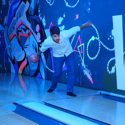 A young man bowls a pink ball down a brightly lit lane with graffiti-style art on the wall.