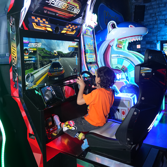 A child plays a racing arcade game, gripping the steering wheel. The game features a car on a winding road.