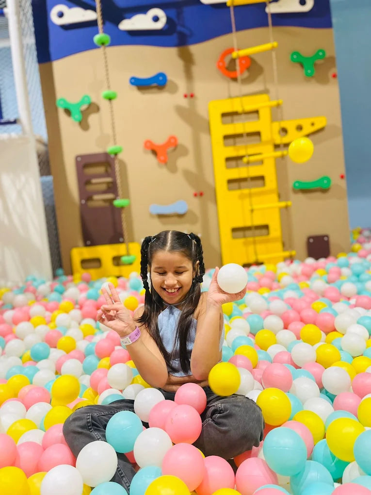 A young girl smiles while holding a white ball in a ball pit, with a climbing wall in the background.