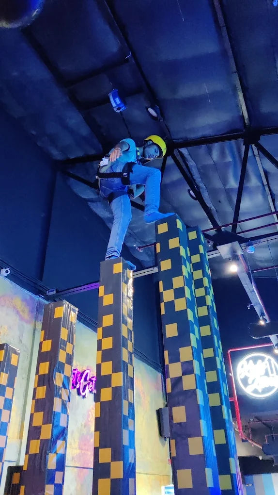 A young person in a harness and helmet stands on top of a tall, checkered pillar in an indoor adventure park.