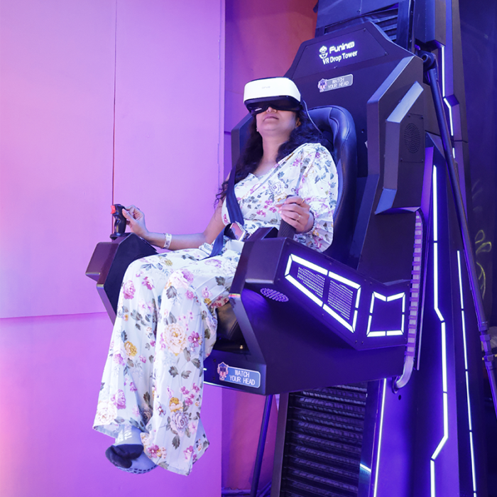 A woman in a VR headset sits in a drop tower ride, holding a joystick. The ride is lit with purple and blue lights.