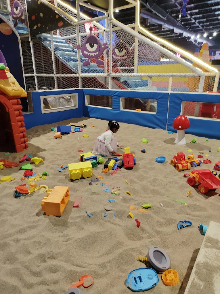 A young child plays in a sandpit filled with toys, surrounded by a colorful indoor playground.