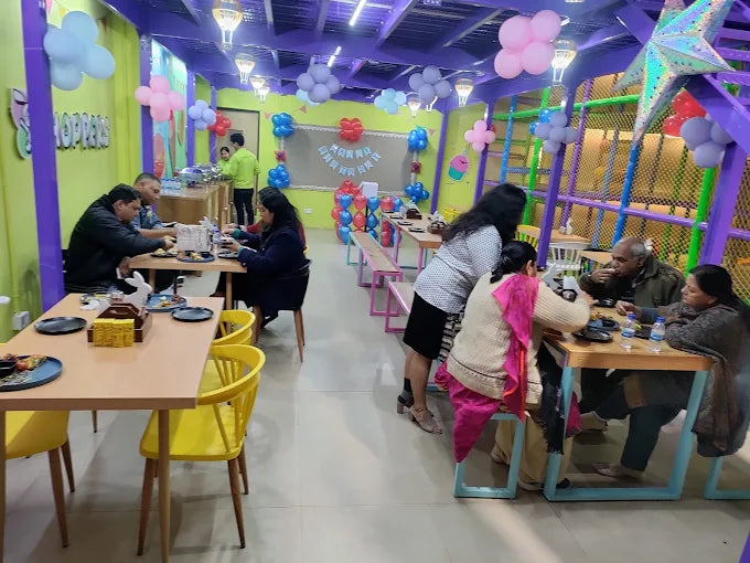 Adults and children enjoying a colorful indoor play area with tables, chairs, balloons, and play structures