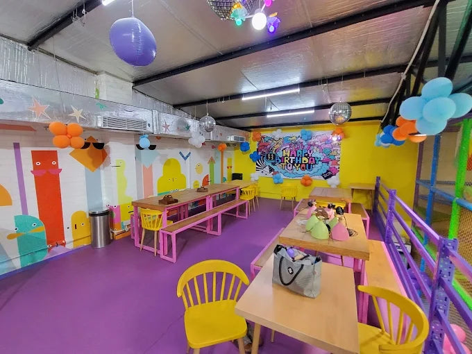 Bright party room with colorful decorations, tables, yellow chairs, and a vibrant birthday banner on the wall