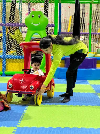 Young child sitting in a red toy car while an adult adjusts the wheels on a colorful soft play mat area