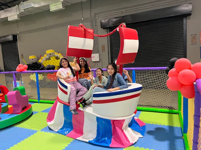Four children sitting on a colorful indoor play structure inside a vibrant padded play area with netted safety barriers