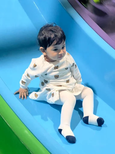 Toddler wearing white outfit sitting at the bottom of a smooth blue slide in a bright play area