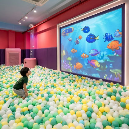A young child stands in a ball pit, looking at a large screen displaying animated fish.