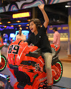A young person joyfully rides a red motorcycle arcade game, waving with excitement.