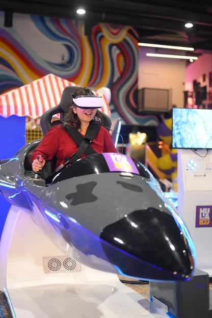 A young woman in a VR headset laughs while operating a flight simulator in an arcade.