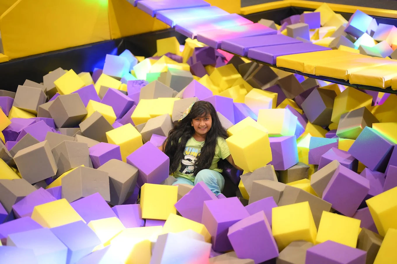 A young girl smiles while sitting in a foam pit filled with colorful cubes.