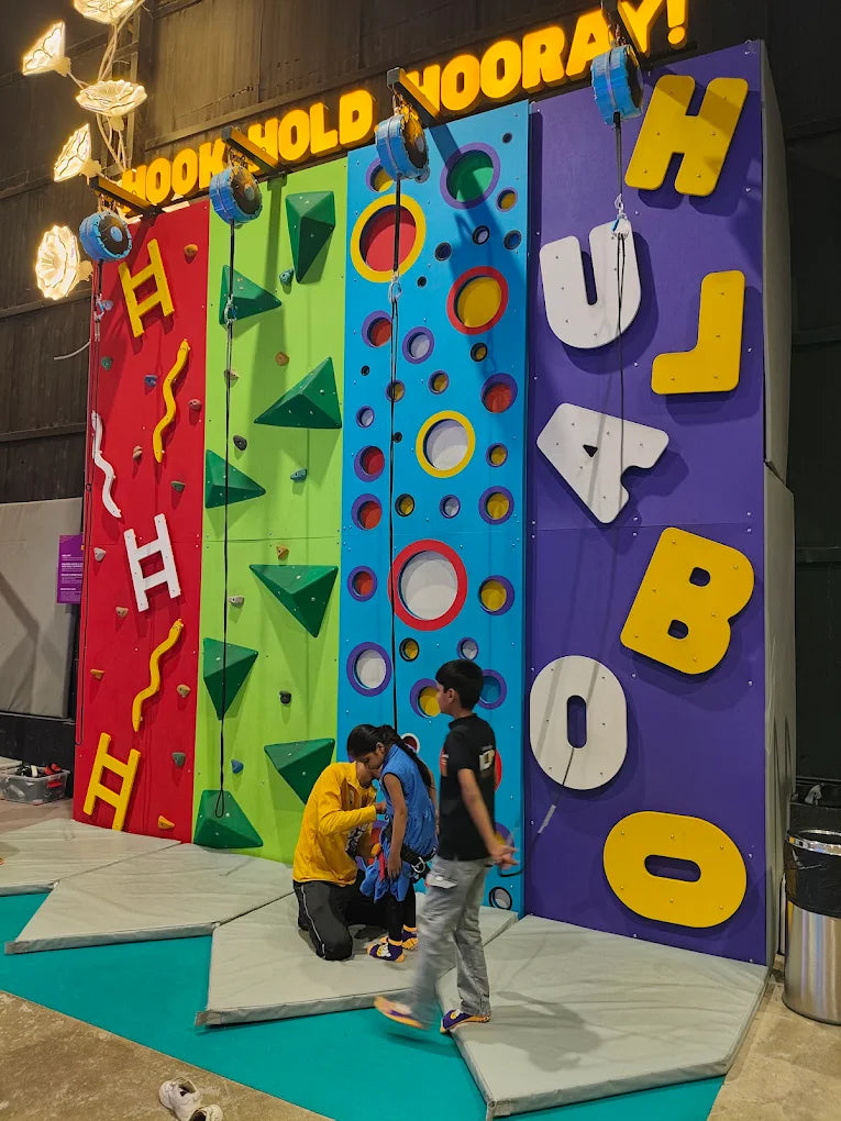Children climb a colorful indoor rock wall with large letters spelling "HOORAY!" on the side.