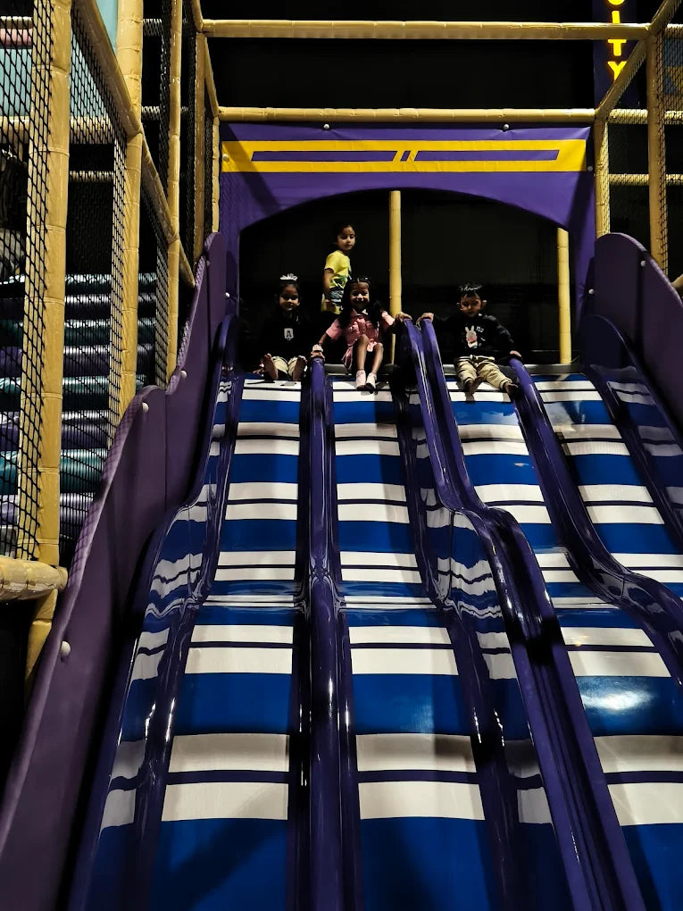 Children sit at the top of a multi-lane blue and white slide in an indoor play area.