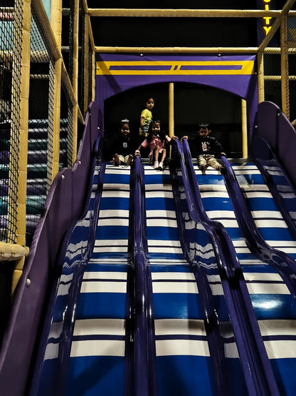 Children sit at the top of a multi-lane blue and white slide in an indoor play area.