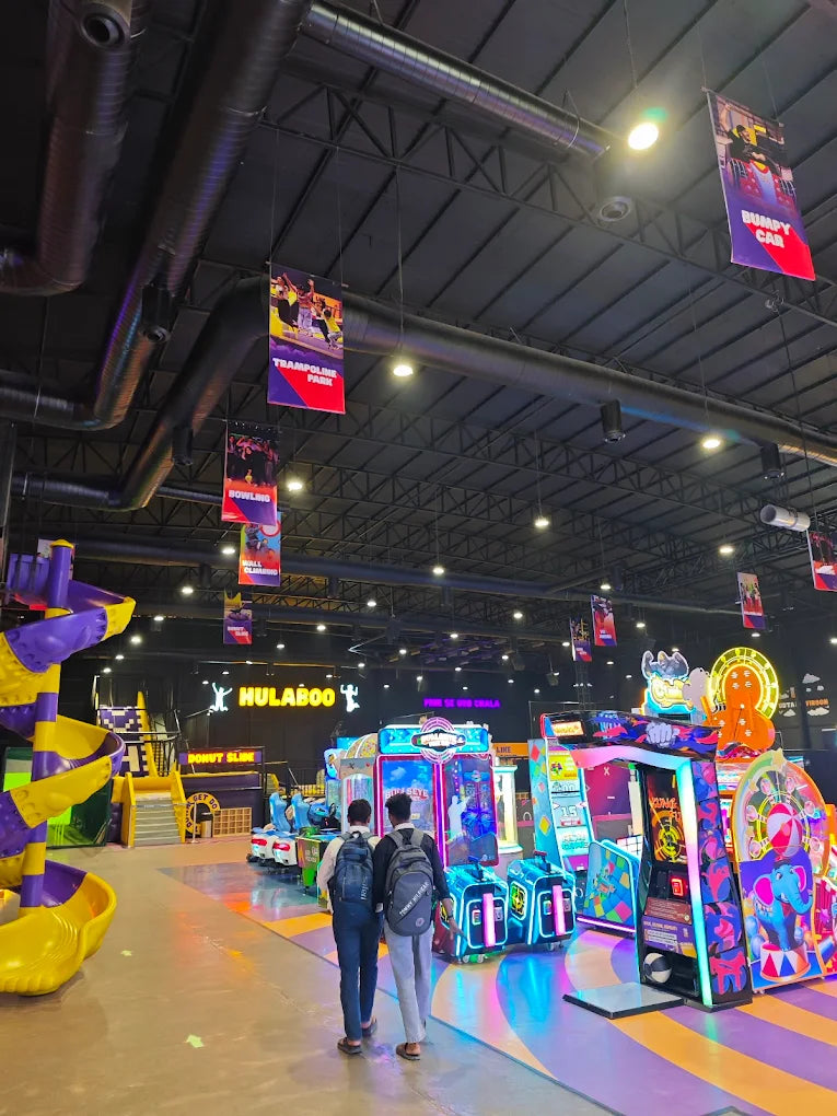 Two young men walk through a brightly lit arcade with colorful games and a large play structure.