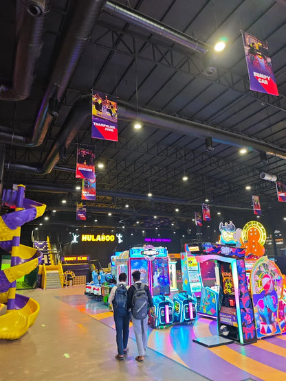 Two young men walk through a brightly lit arcade with colorful games and a large play structure.