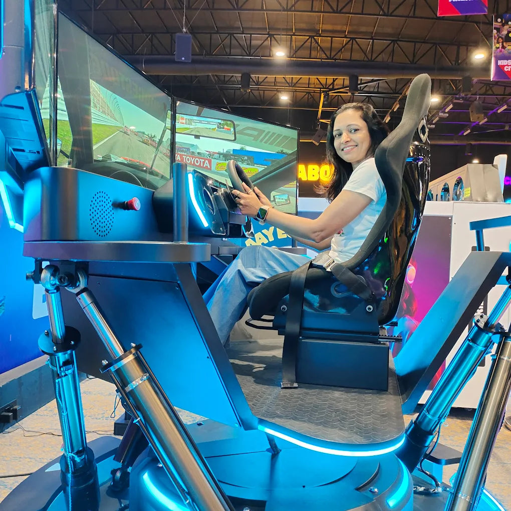 A woman sits in a racing simulator, holding the steering wheel and smiling at the camera. The simulator has multiple screens displaying a race track.