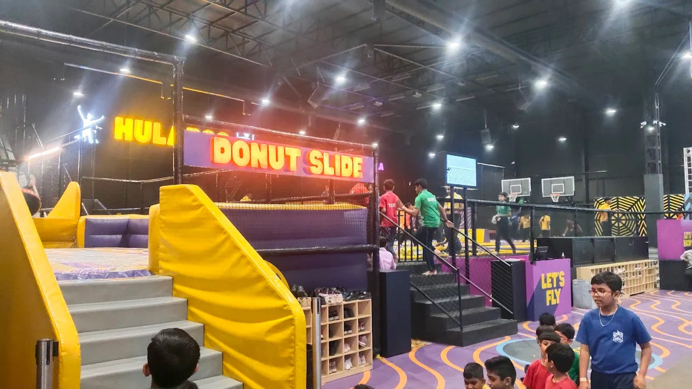 Children and staff at an indoor trampoline park with a "Donut Slide" and basketball hoops.