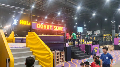 Children and staff at an indoor trampoline park with a "Donut Slide" and basketball hoops.