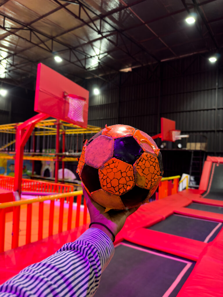 A person holds a soccer ball in a trampoline park with red and orange trampolines.