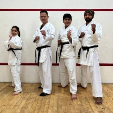 Four martial artists in white uniforms with black belts posing in a room with wooden flooring and white walls.