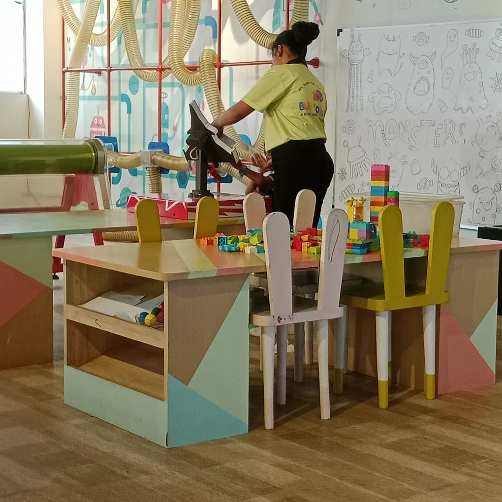 Children's play area with colorful tables, chairs, and building blocks, while a woman supervises.