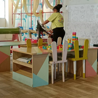 Children's play area with colorful tables, chairs, and building blocks, while a woman supervises.