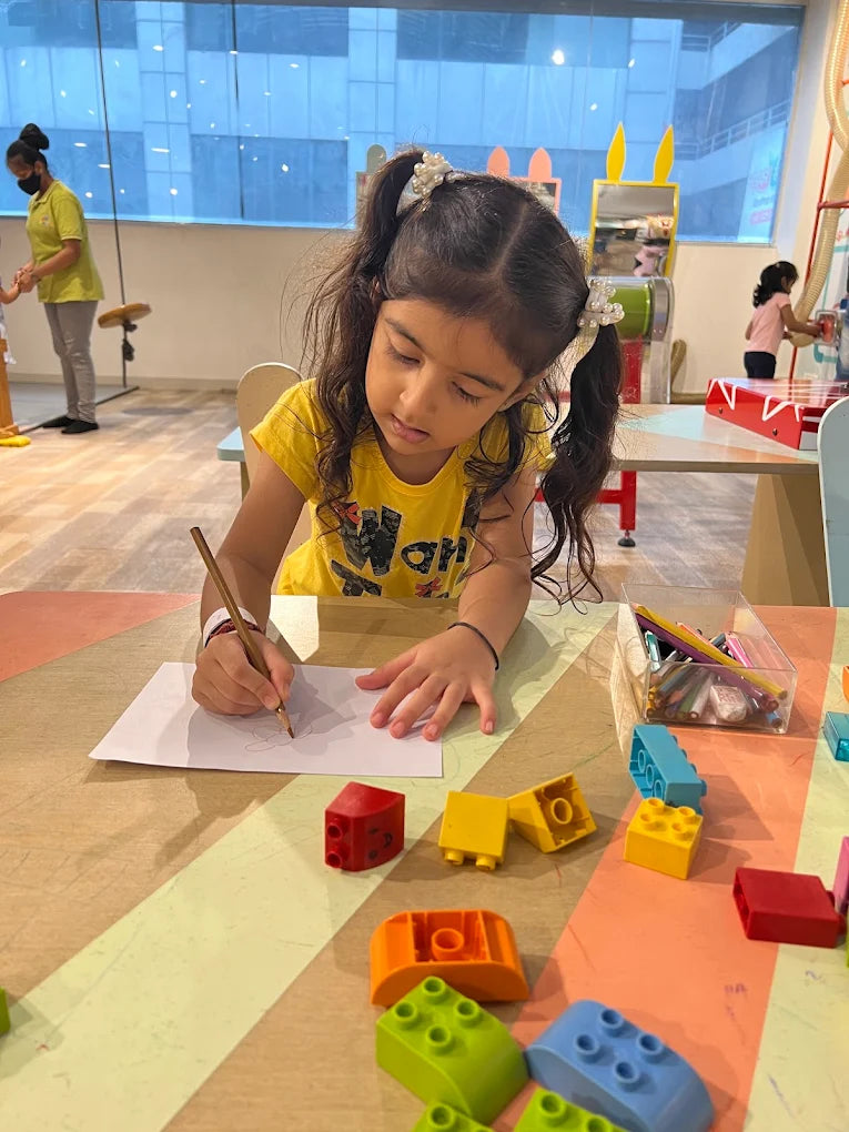 A young girl draws at a table with colorful building blocks scattered around her.