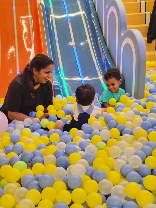 A woman and two young children play in a ball pit with a slide in the background.