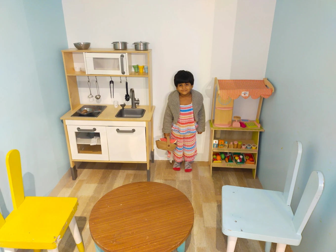 A young child stands in a play area with a toy kitchen and a small market stall.