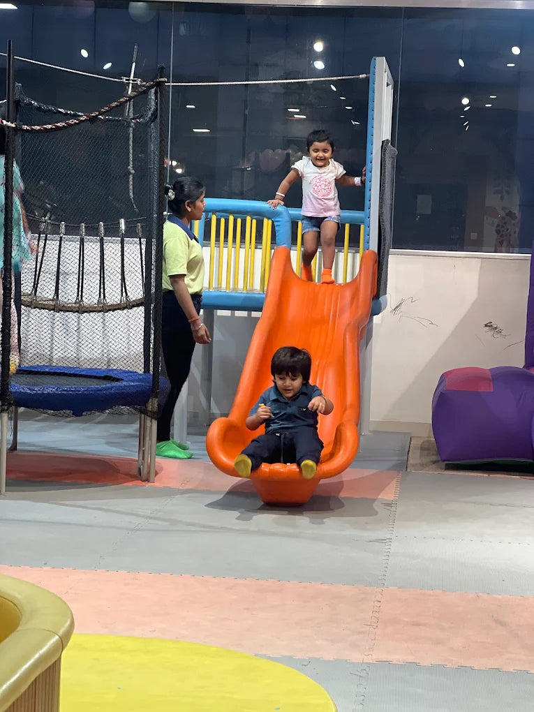 A young child slides down an orange slide while another child stands at the top, supervised by an adult.