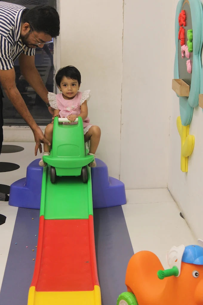 A man helps a young child on a toy car slide in a playroom.