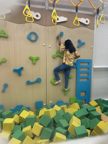 A young child climbs a colorful indoor climbing wall above a pit of foam blocks.