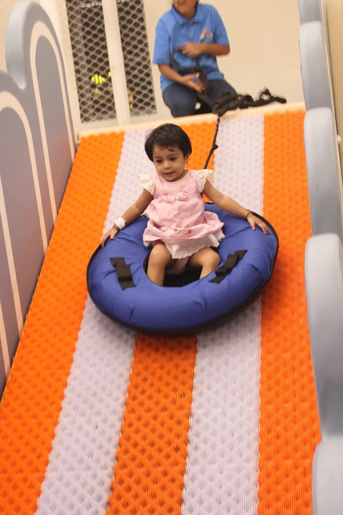 A toddler slides down a colorful ramp in a blue tube, pulled by an adult.